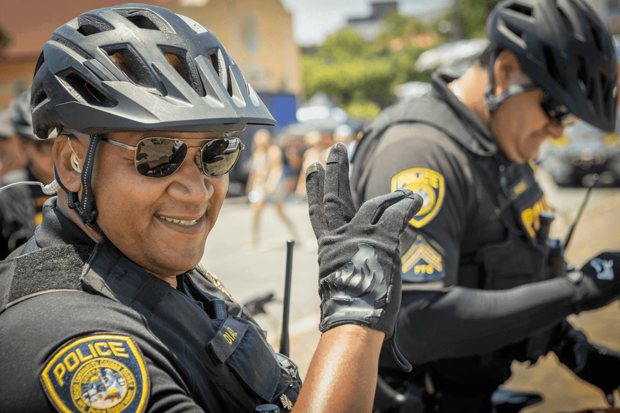 A college police officer wearing gloves and a bicycle helmet waves as he patrols a parade on a bike.