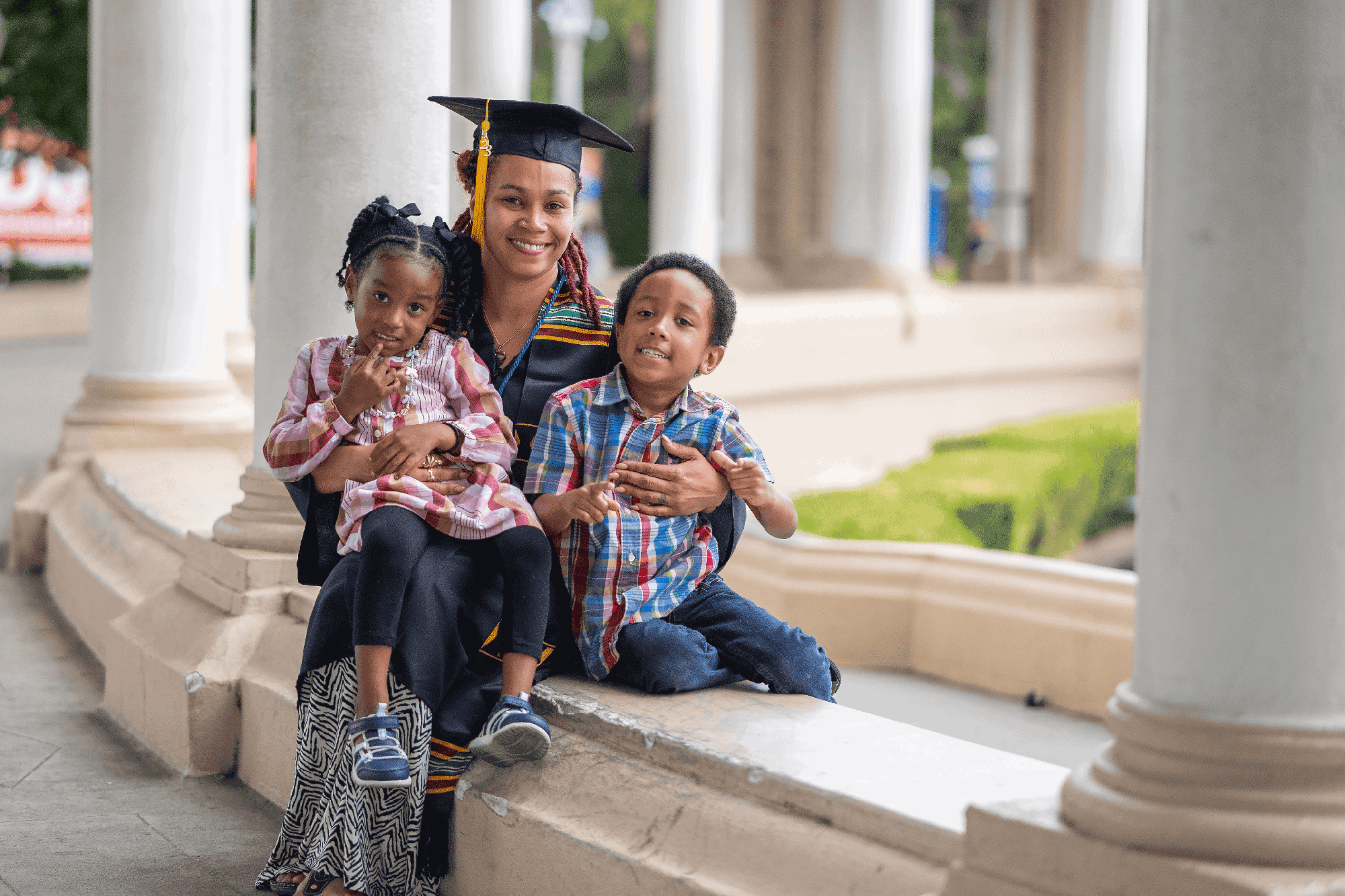A mother in a graduation cap and gown holds her two children