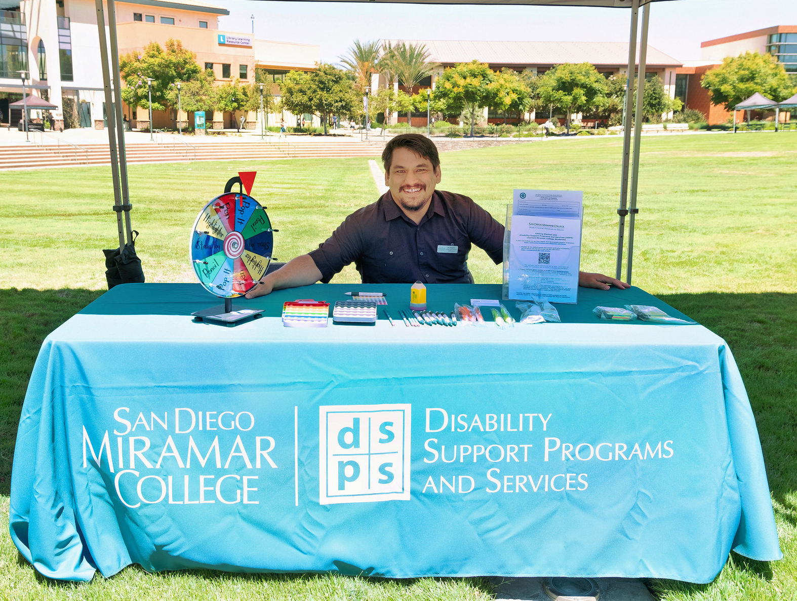 A man sitting at a welcome booth with a prize wheel that people can spin and win a prize.