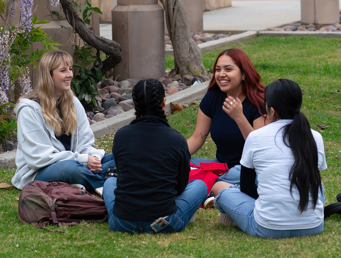 Four women sitting on the grass with backpacks talking and laughing.