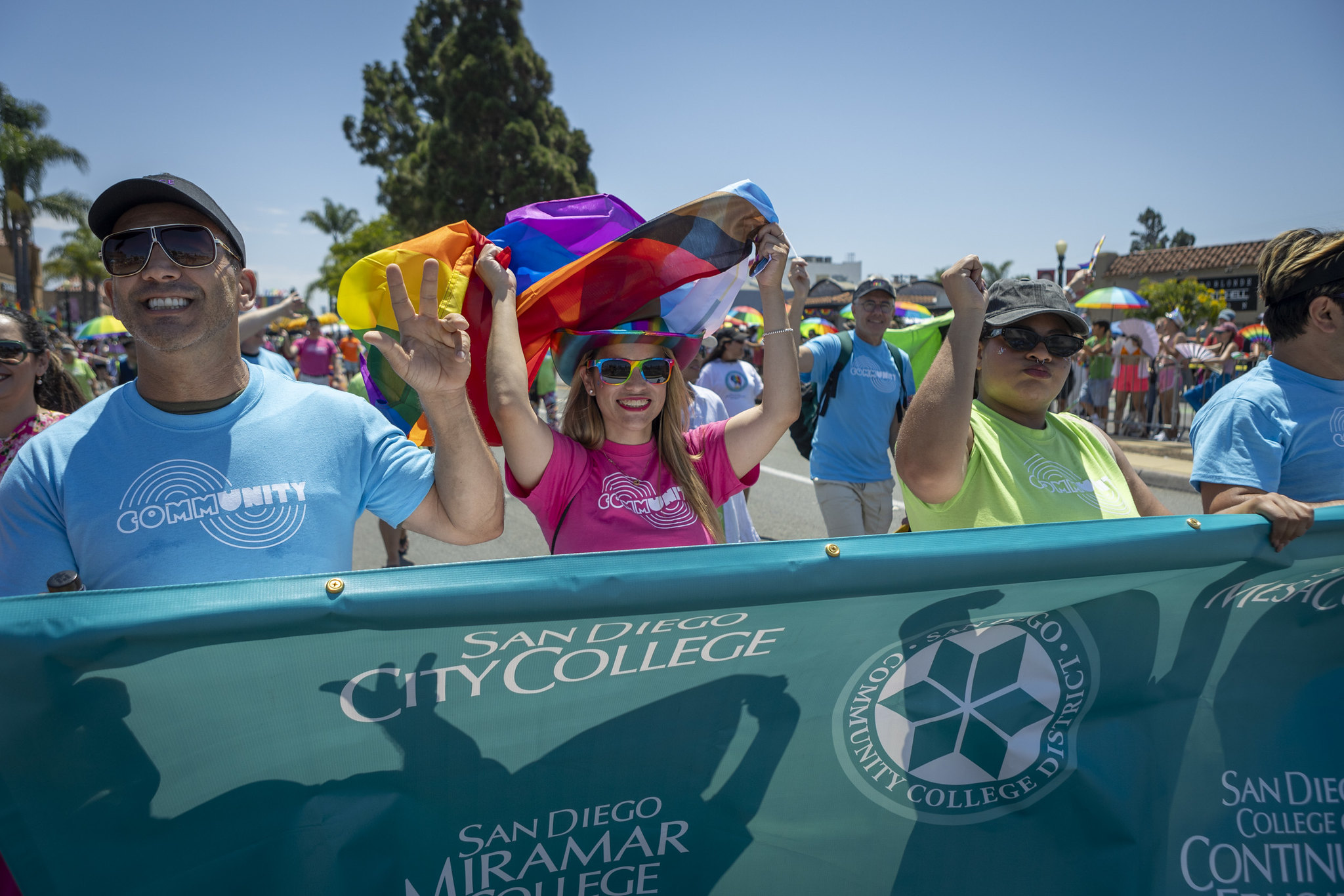 Four people wearing matching tee shirts that say community, one green, one pink and two blue. There are holding a banner and getting ready to march in the Pride Parade. One lady is waving a rainbow flag.