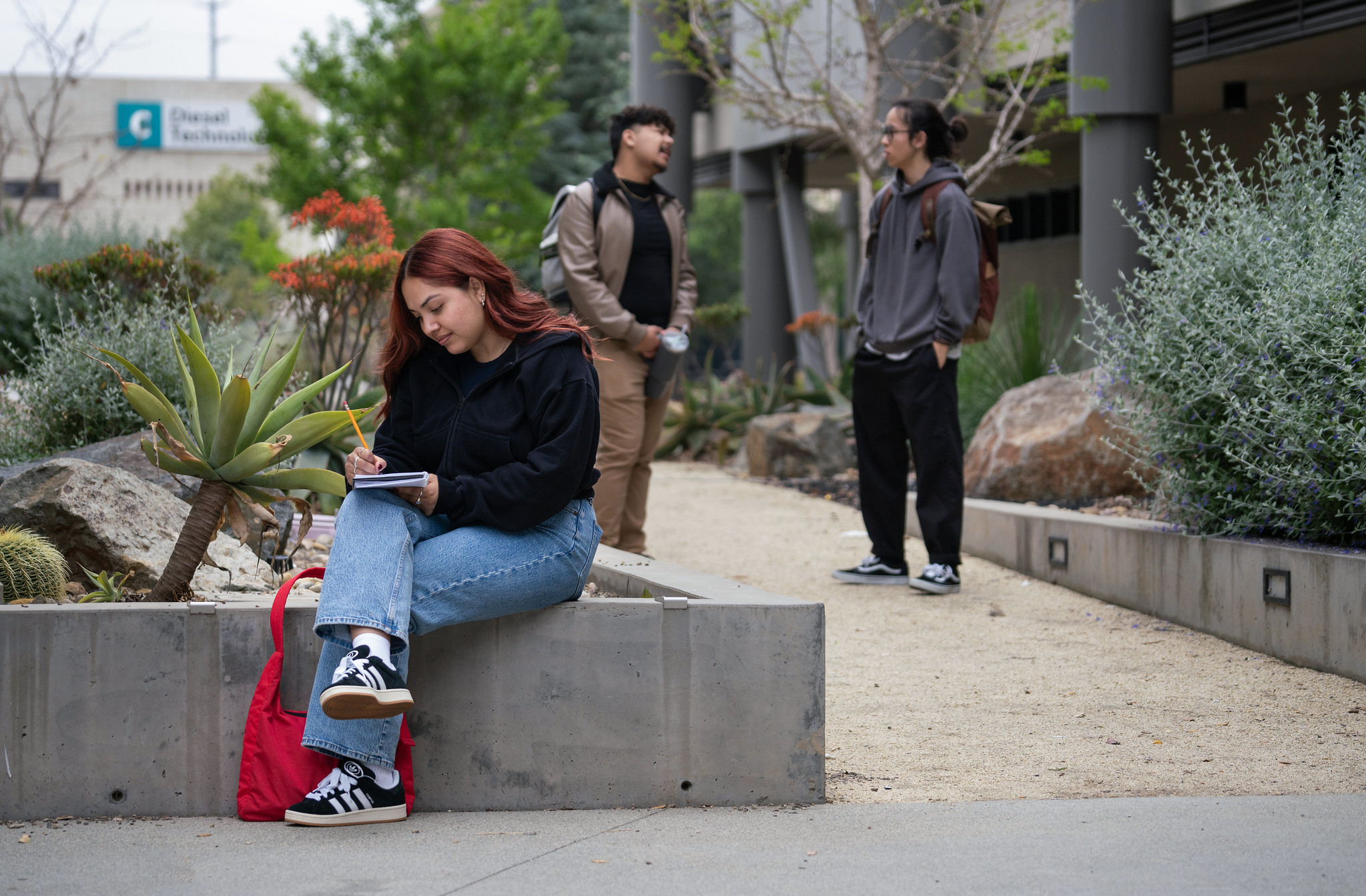 Three students outside at Miramar campus. Two are talking and one is seated writing in a notebook