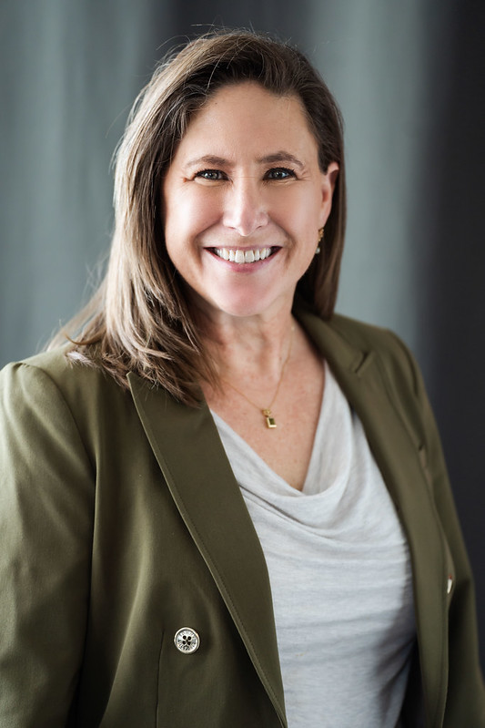 Alison, a white woman with shoulder-length light brown hair, smiles at the camera wearing a light bue draped shirt and green blazer
