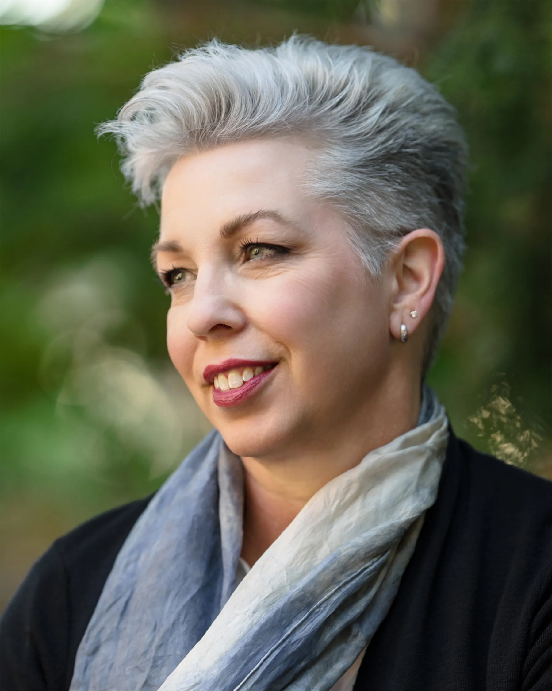 Image of Flower Darby, a white woman with short, white hair wearing a black shirt and gray scarf smiling, looking off camera