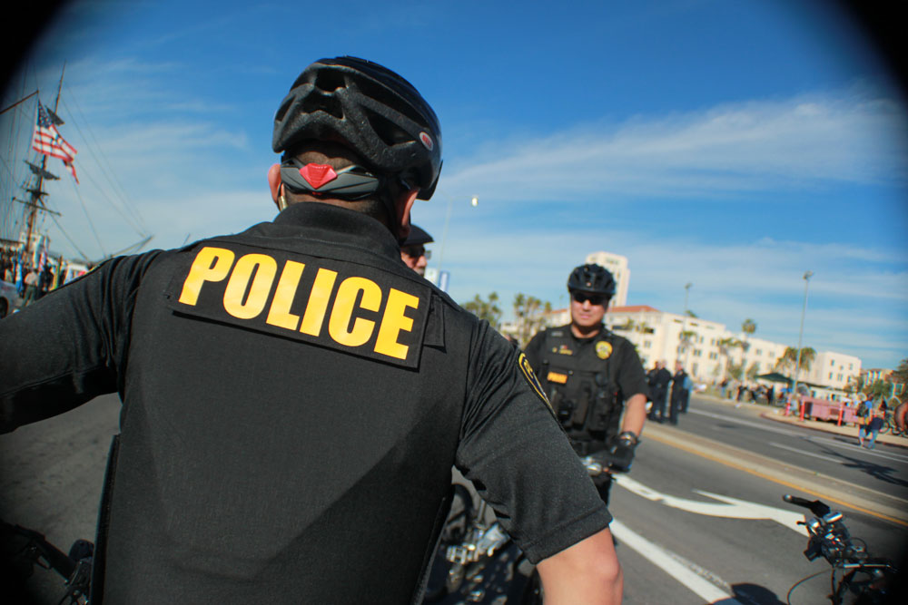 The back of an officer. His black button down shirt has the word police in yellow
