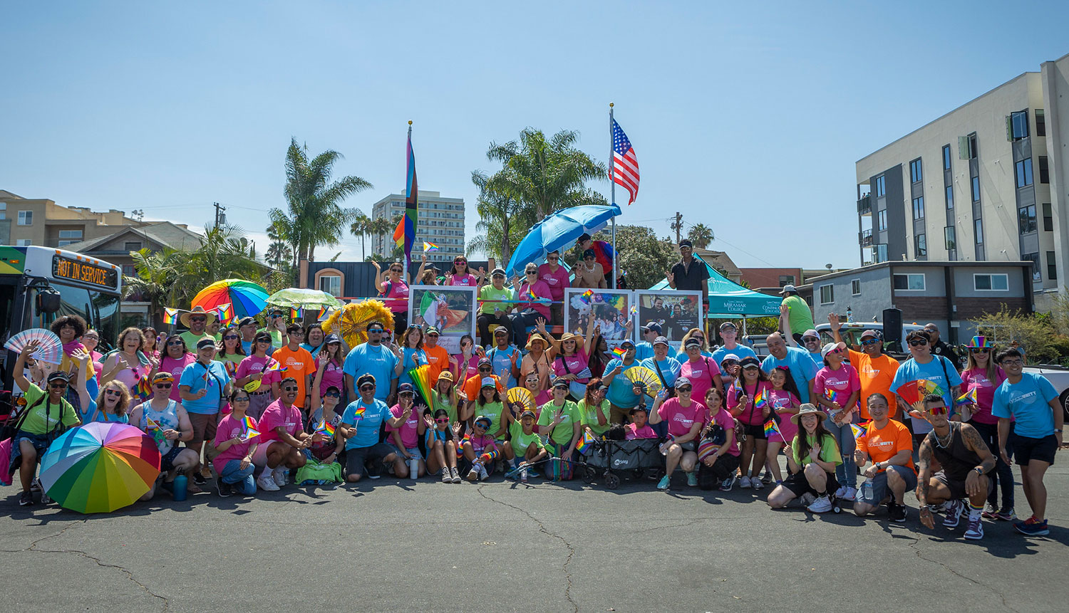 A group photo of employees and staff at the pride parade they are all wearing matching tee shirts that say community. Each shirt is green, blue, pink or orange.