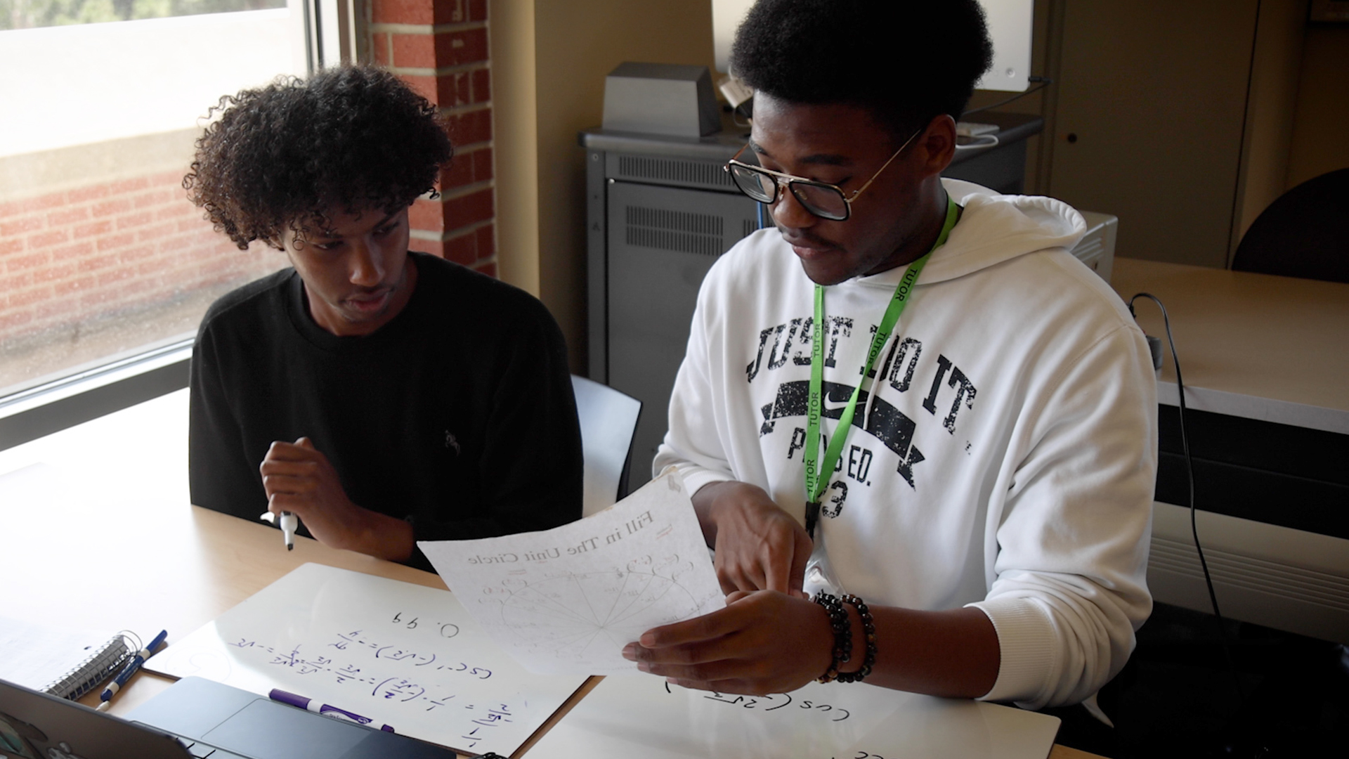 A tutor is showing a student a piece of paper while they're sitting at a desk