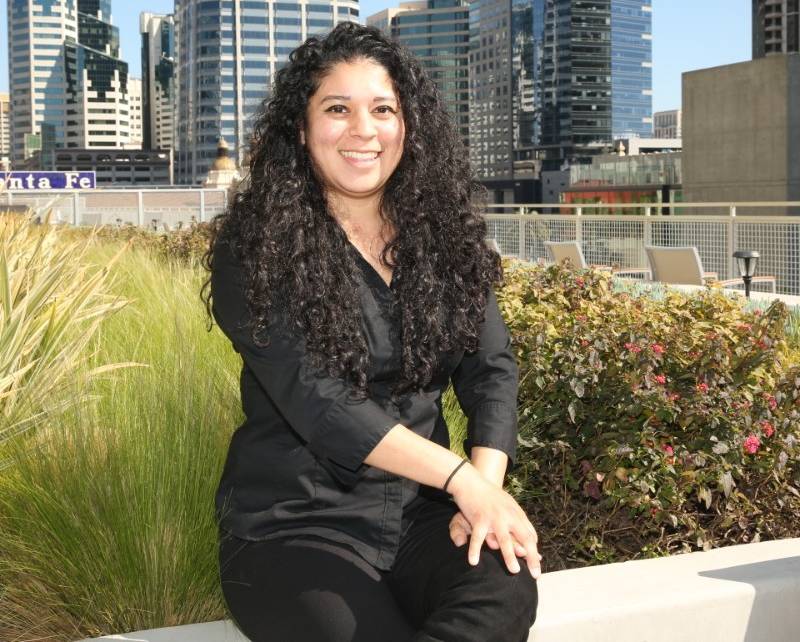 A smiling Jessica Ramos-Bahena, wearing a black shirt and pants, is sitting outside in front of a scenic downtown San Diego background.