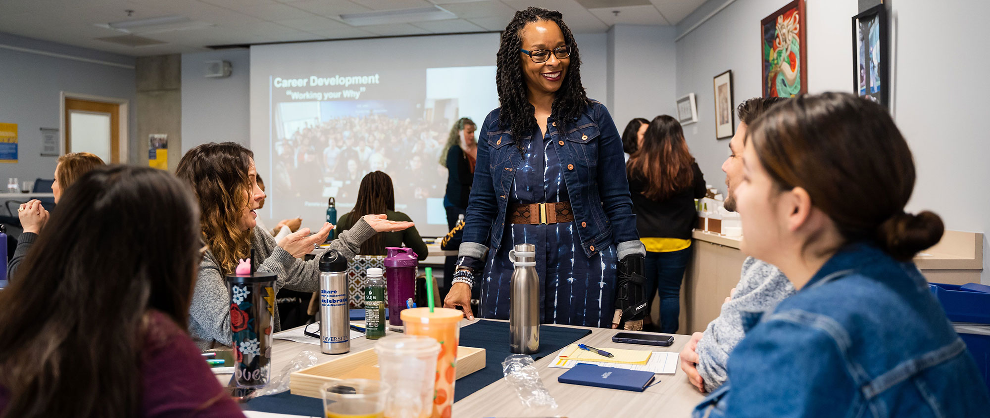 Mesa College President Ashanti Hands is smiling and standing while speaking to three students sitting at a table in a classroom.