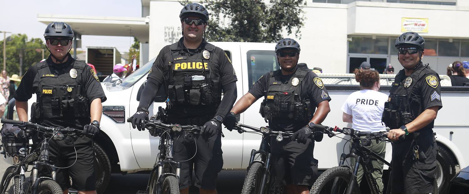 Four college officers on bike patrol are wearing helmets and standing next to their bicycles.