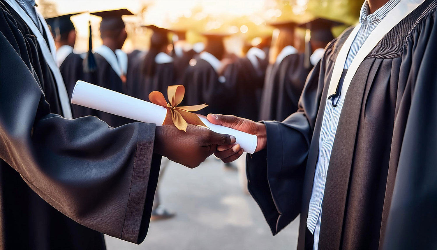 A closeup of two hands. One is handing the other a graduation certificate with a gold bow. Both are wearing black graduation gowns.