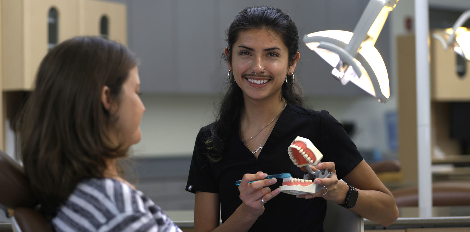 Anthony Ramirez is wearing black scrubs and holding a plastic set of teeth and a toothbrush. A lady is next to him sitting in a dentist chair