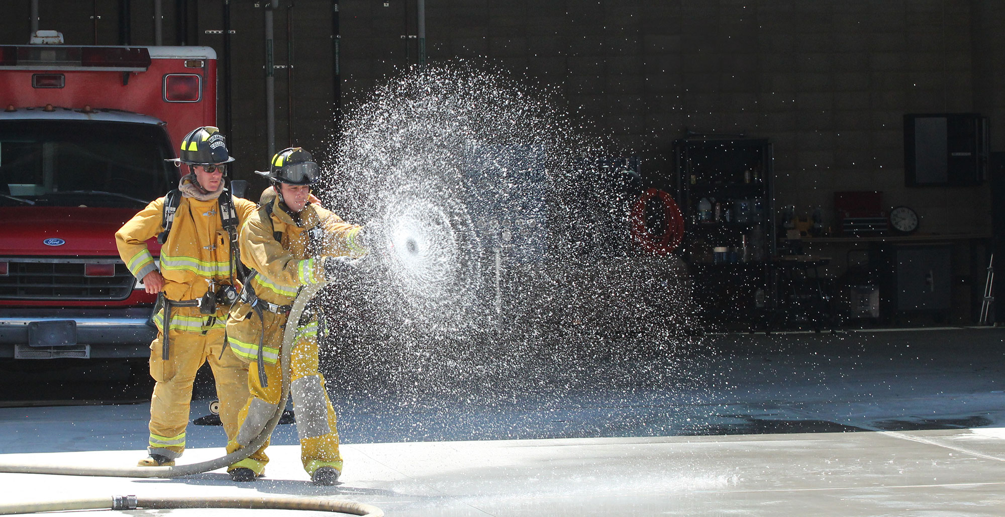 Two fire academy students in yellow fire gear use a fire hose. The water from the hose is making a spiral