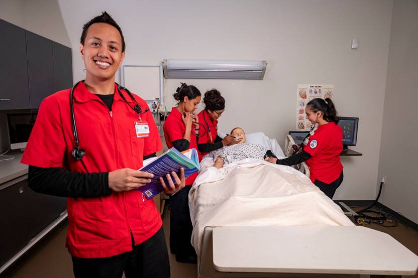Nursing students Four nursing students wearing red and black scrubs check on a dummy patient in a hospital bed