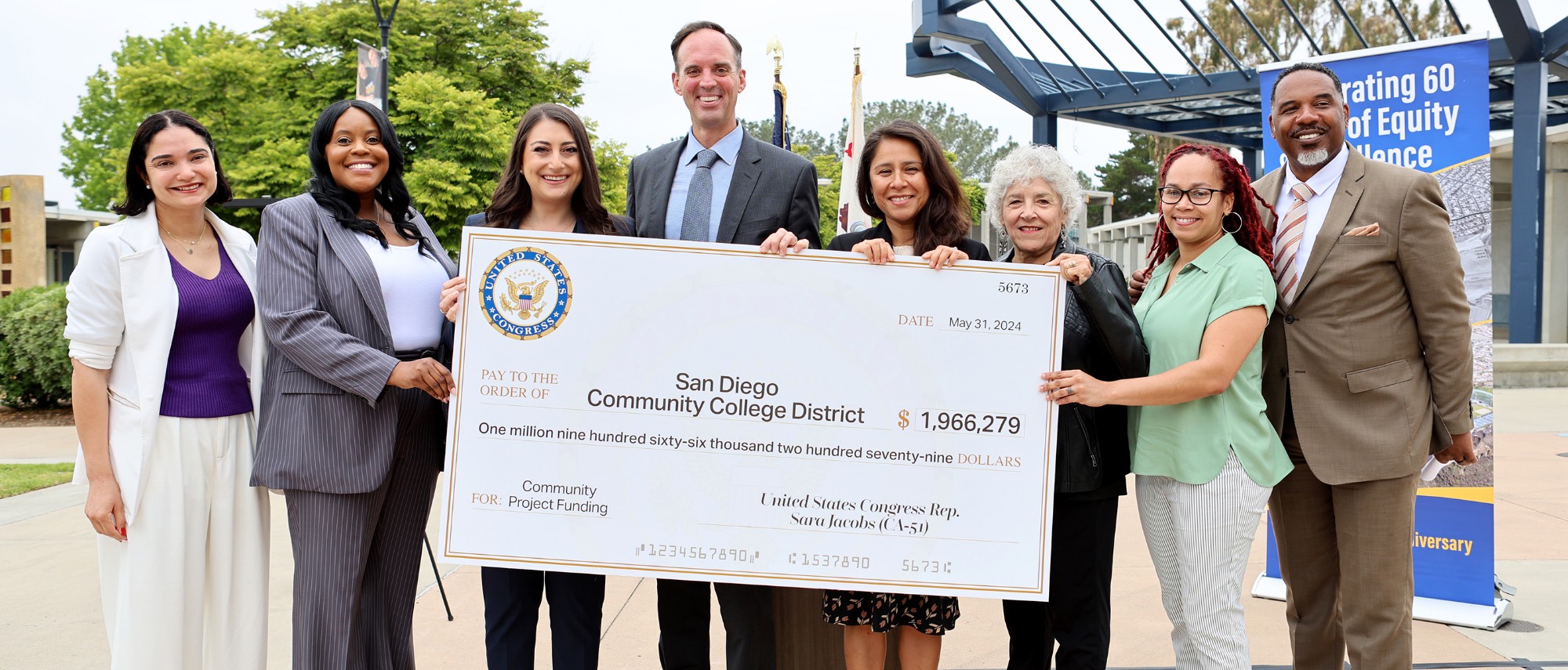 Eight people hold up a giant check for nearly two million dollars. They are outside in a courtyard.