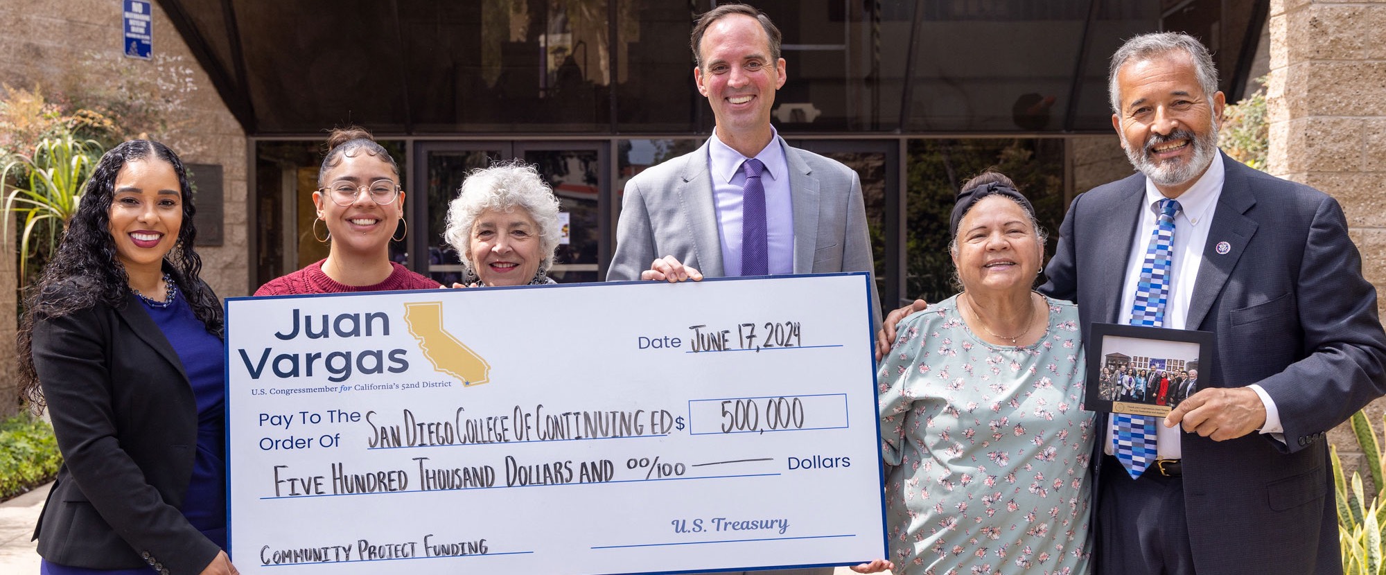 Six people hold up a giant check for 500,000 dollars outside of the entrance of the Educational Cultural Complex