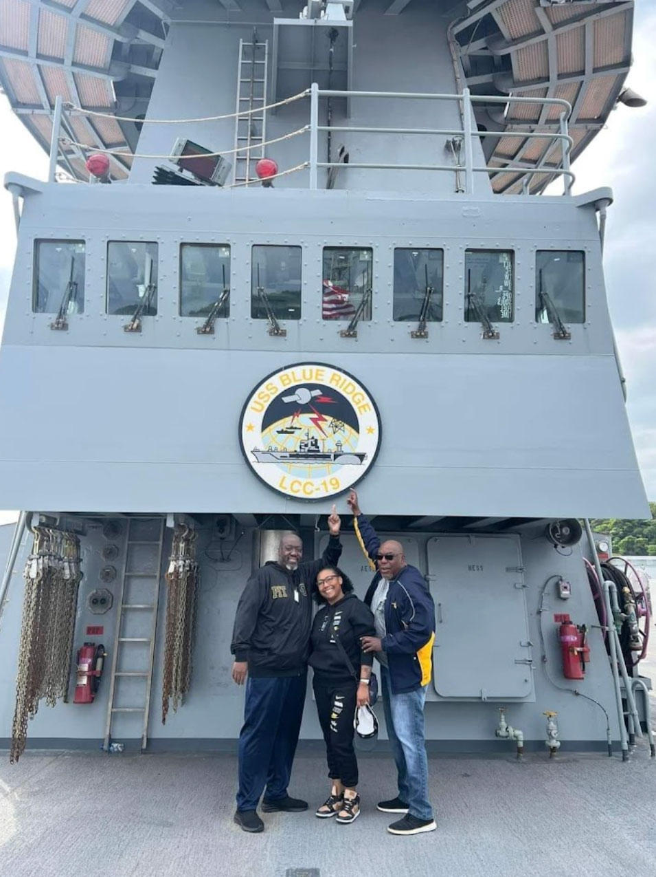 Three people stand in front of the bridge of a Navy ship
