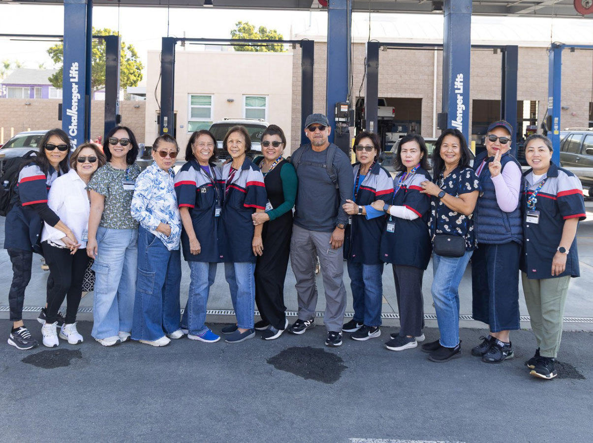 13 people in front of the automotive work area at continuing education. Six are wearing blue and red mechanic shirts.