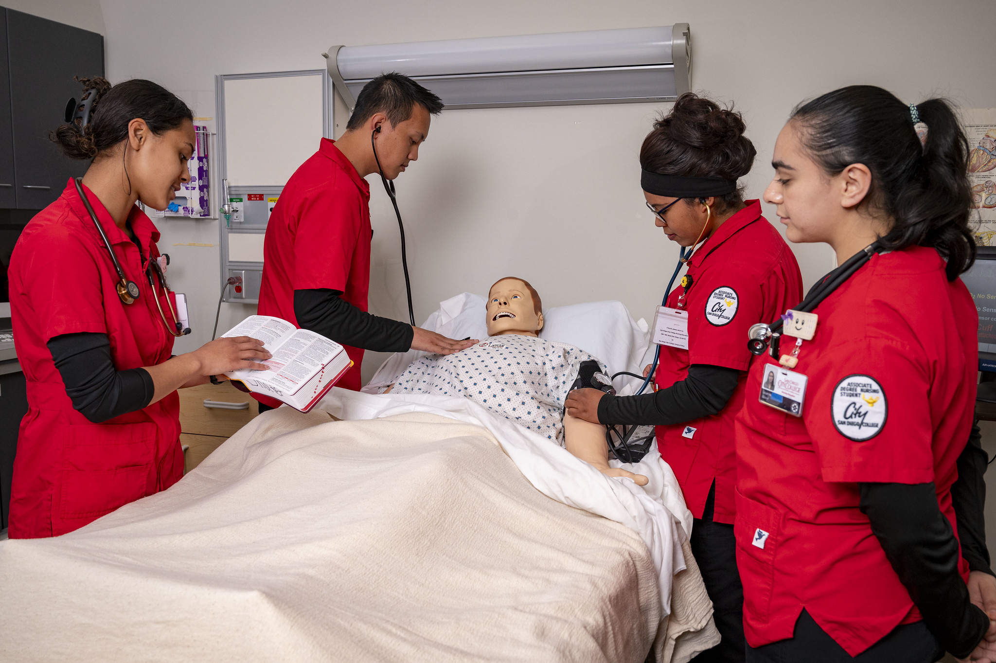 Four students in red smocks stand around a bed with a dummy patient
