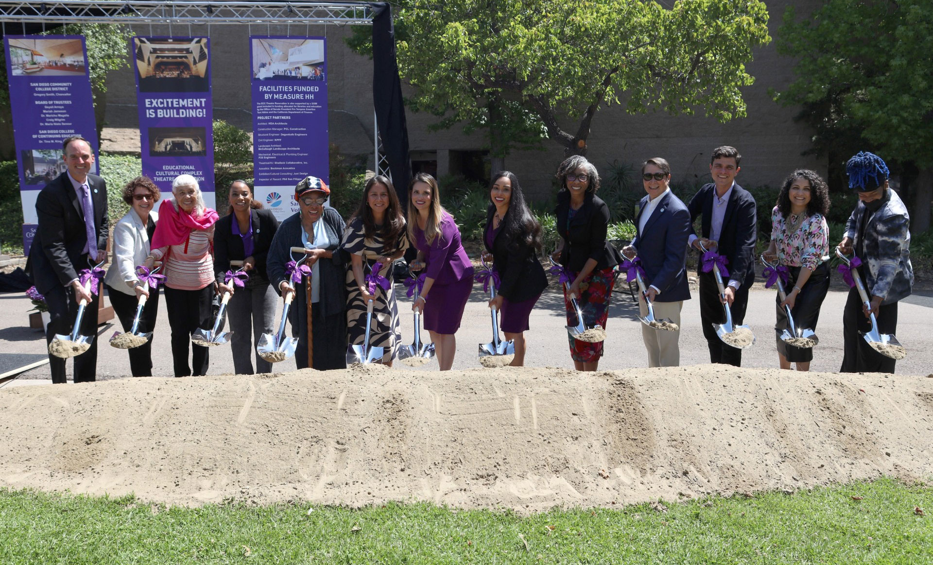 Thirteen people lined up shoveling dirt at a groundbreaking ceremony.