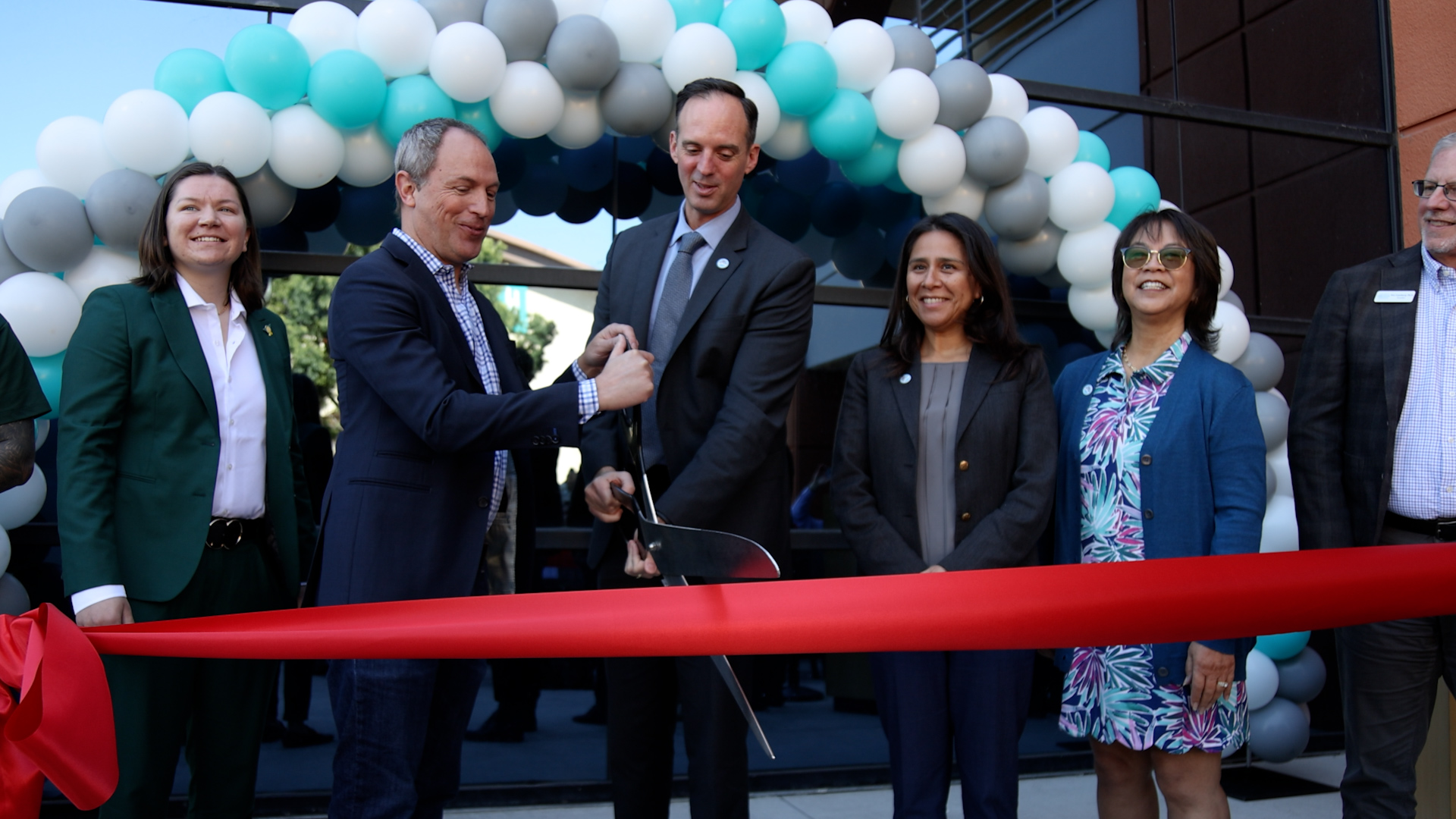 Two people holding giant scissors cutting a red ribbon.