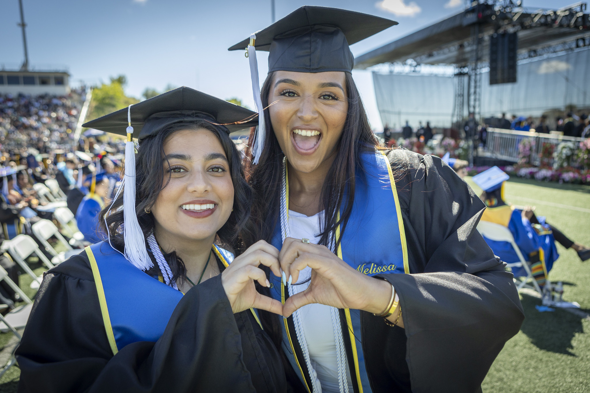Two students wearing graduation caps and gowns at commencement put their hands together to make a heart.