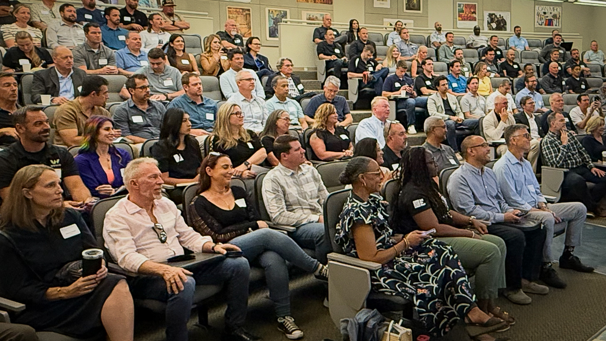 A large room with slight stadium seating almost completely filled with men and women seated and watching a presentation happening at the front of the room, which includes one female talking and a project showing a rendering of the building project being discussed. 