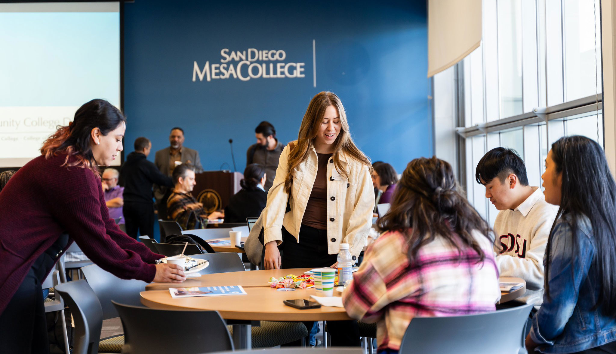Five students sit and stand around a table. There is a blue wall that says San Diego Mesa College